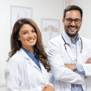 Uma fotográfia, uma médica e um médico sorrindo, ambos com jalécos. A médica está na faixa dos 30 anos, tem o cabelo castanho e uma blusa jeans azul por baixo do jaléco. O médico também na faixa dos 30, está com braços cruzados.